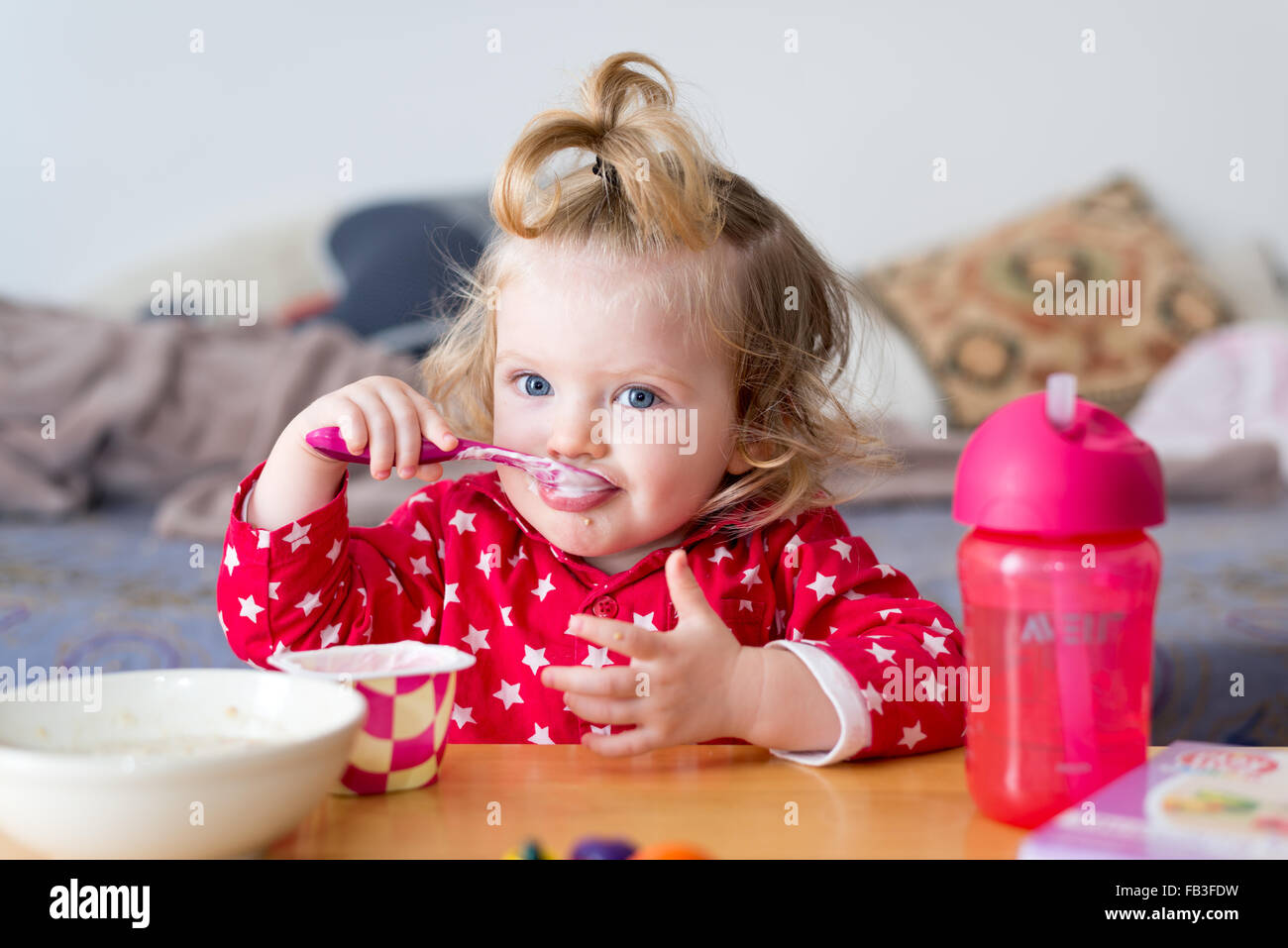 18 mese Baby girl mangiare yogurt da se stessa con un cucchiaio Foto Stock