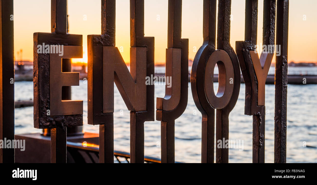 La parola "godere' inset in ringhiere in ferro presso il World Financial Plaza con un tramonto sul fiume Hudson dietro Foto Stock