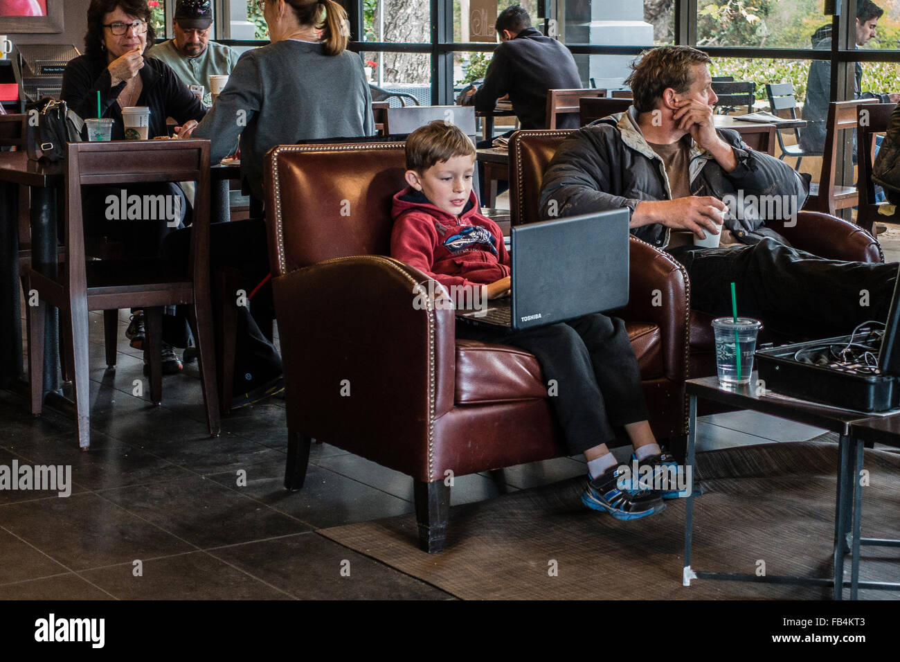 Un giovane tecnicamente savvy boy, 10-12 anni, si siede in una caffetteria con un grande computer portatile sul suo giro come egli si concentra su di esso. Foto Stock