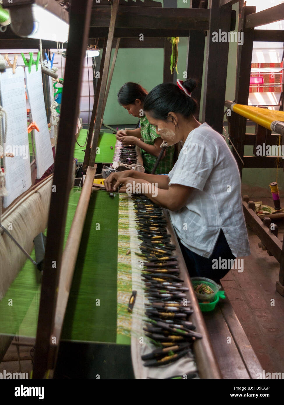 Due donne tessitura utilizzando navette con bobine di filo su di un telaio di tessitura. Parzialmente in tessuto di seta verde visibile del tessuto. Mandalay, Myanmar. Foto Stock