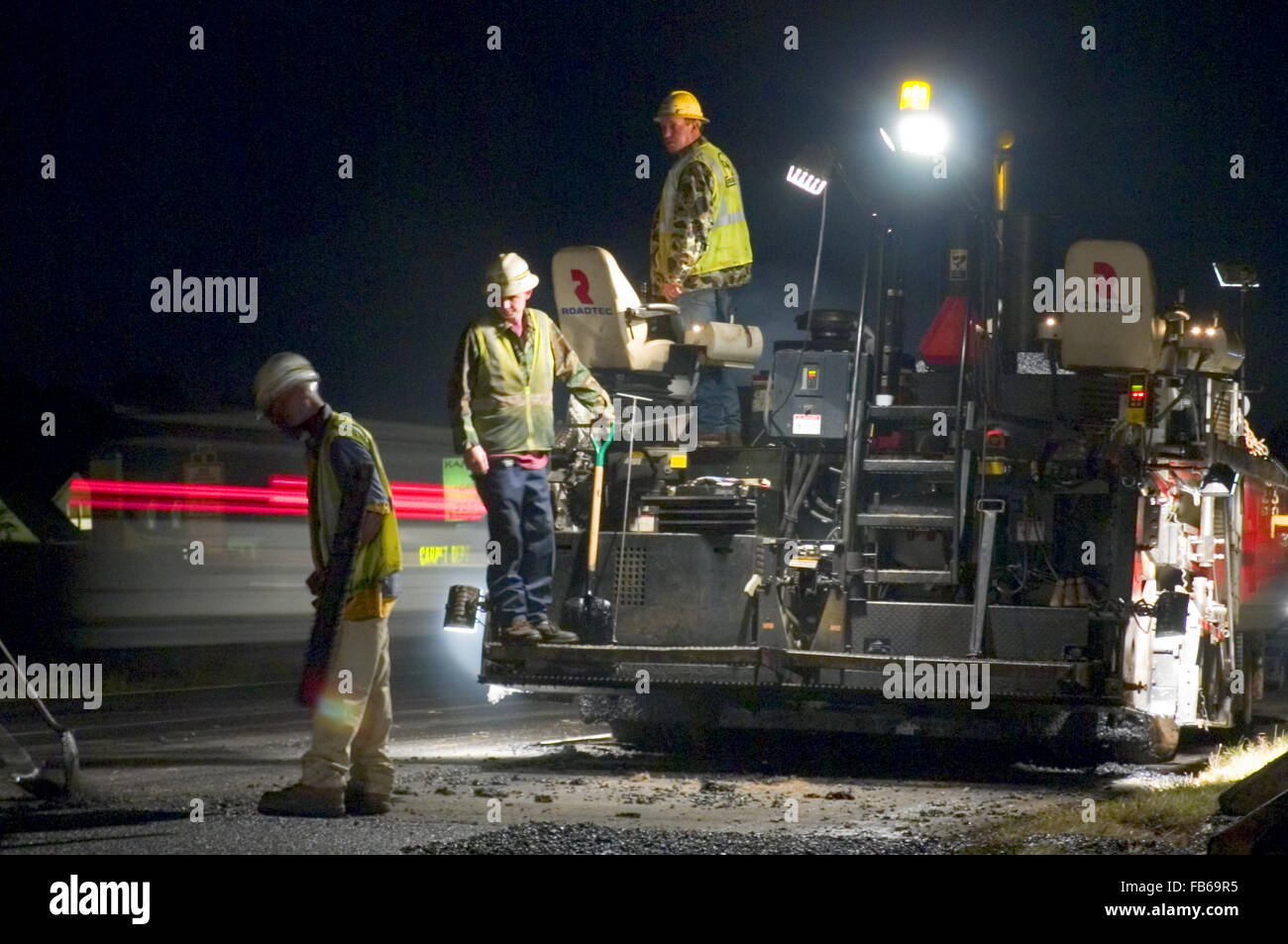 I lavoratori di sostare dietro un asfalto spandiconcime immissione hot mix durante la strada interstatale di lavorare di notte. Foto Stock