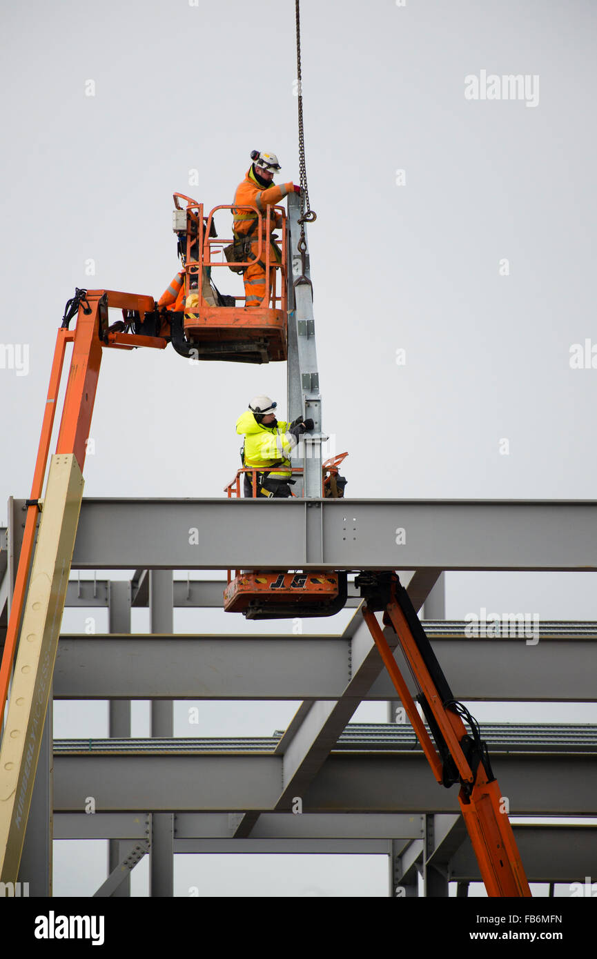 I lavori di costruzione nel Regno Unito : uomini al lavoro in altezza su elevato 'cherry picker' piattaforme, imbullonando insieme il telaio di acciaio di un nuovo sviluppo per il supermercato Tesco e Marks & Spencer store su un sito di costruzione in Aberystwyth, Wales UK Foto Stock