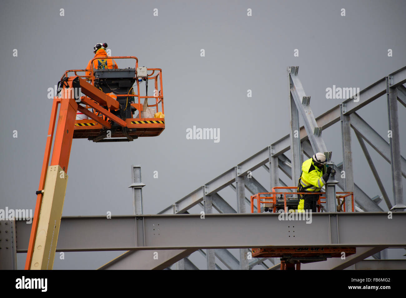 I lavori di costruzione nel Regno Unito : uomini al lavoro in altezza su elevato 'cherry picker' piattaforme, imbullonando insieme il telaio di acciaio di un nuovo sviluppo per il supermercato Tesco e Marks & Spencer store su un sito di costruzione in Aberystwyth, Wales UK Foto Stock