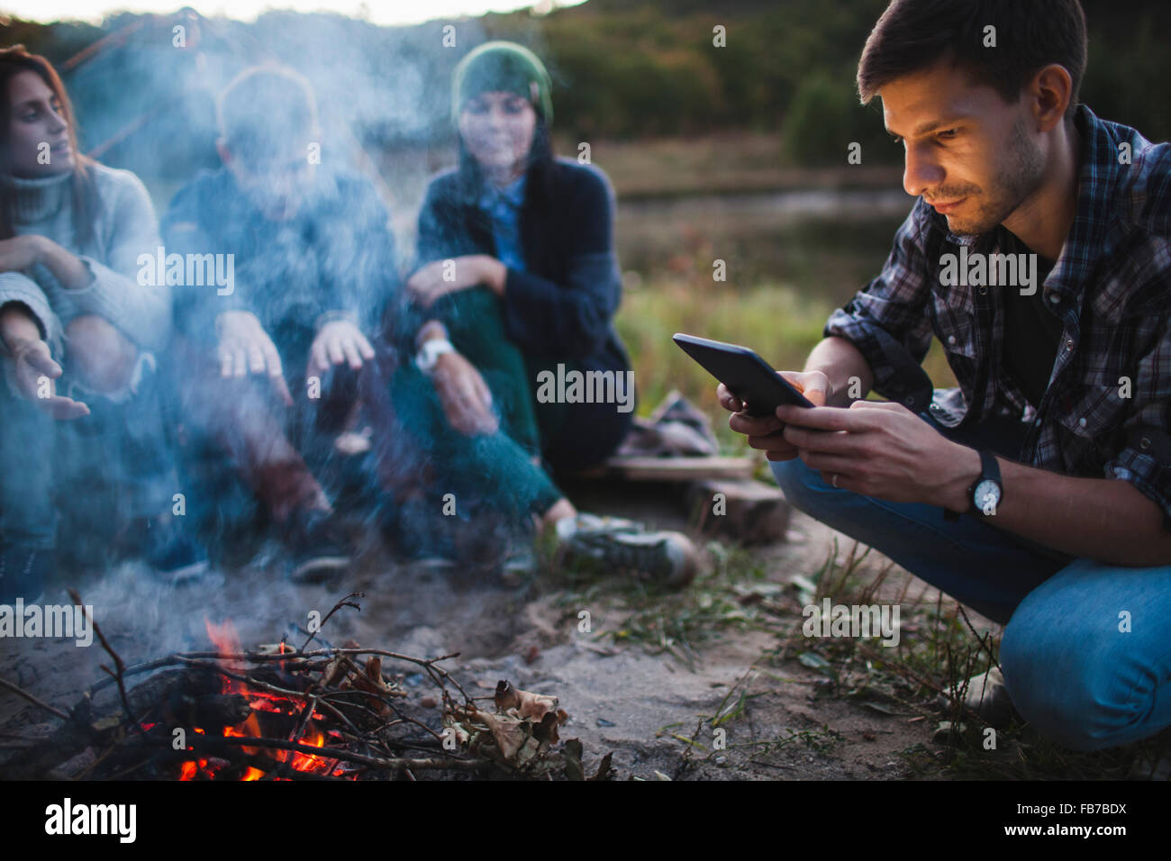 Giovane uomo con tavoletta digitale mentre si siede con gli amici dai falò Foto Stock