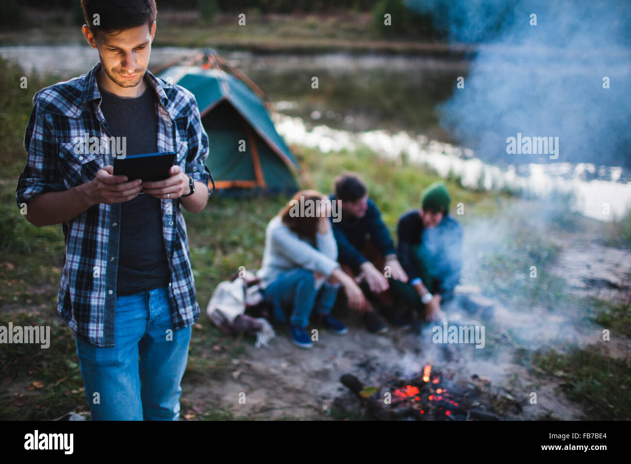 Giovane uomo con tavoletta digitale mentre gli amici seduti davanti al falò in foresta Foto Stock