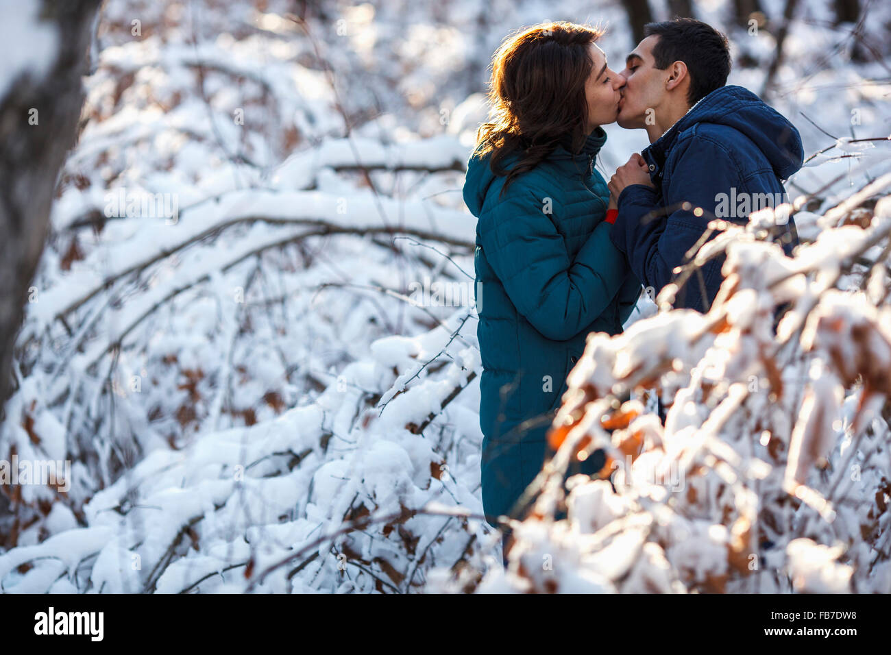 Vista laterale della coppia giovane baciare dalla coperta di neve alberi Foto Stock