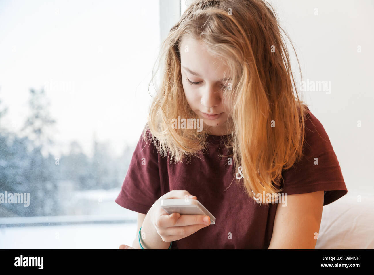 La bionda ragazza seduta vicino alla finestra invernale con lo smartphone Foto Stock