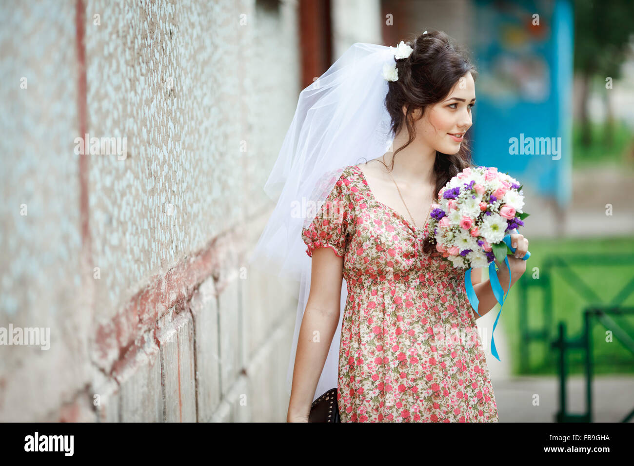 La sposa in semplice abito retrò con motivo floreale, già indossa velo, bouquet nuziali e borsetta, ponendo al di fuori di casa, guardando al lato. Foto Stock