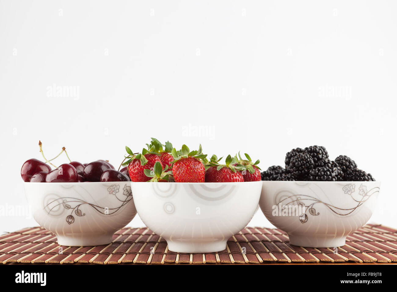 Tre tazze di ceramica con frutti di bosco - fragole, more e ciliegie sul tappetino di bambù isolato su bianco Foto Stock