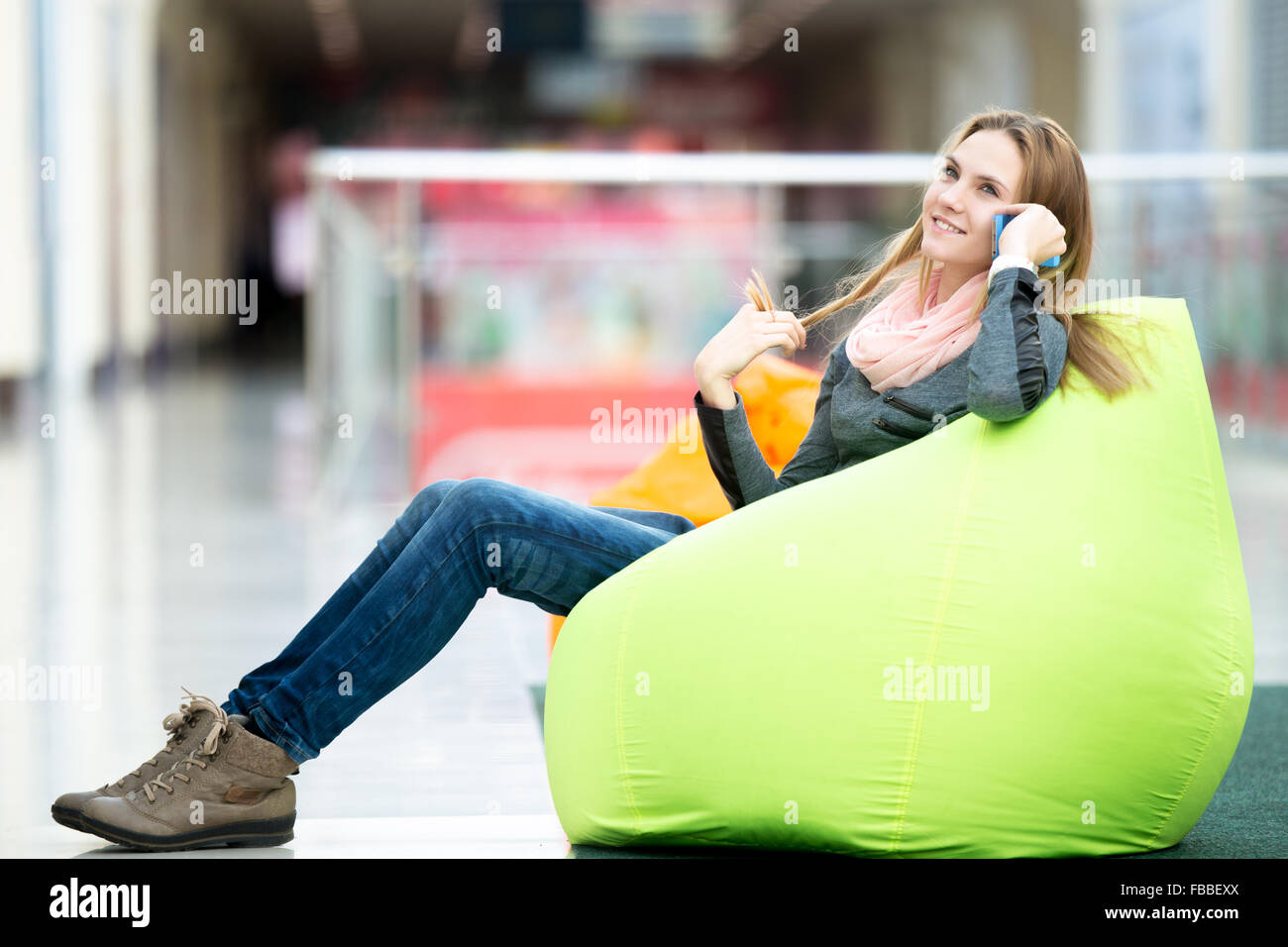 Dreamily sorridente giovane donna in stile casual vestiti seduto sul bean bag in interni contemporanei, area ricreativa, utilizzando smar Foto Stock