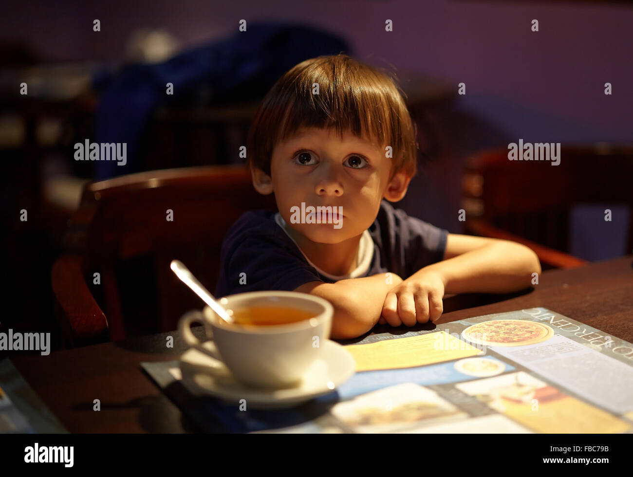 Il bambino in una caffetteria con una tazza,seduto con le mani piegate,grandi occhi,cercando Foto Stock