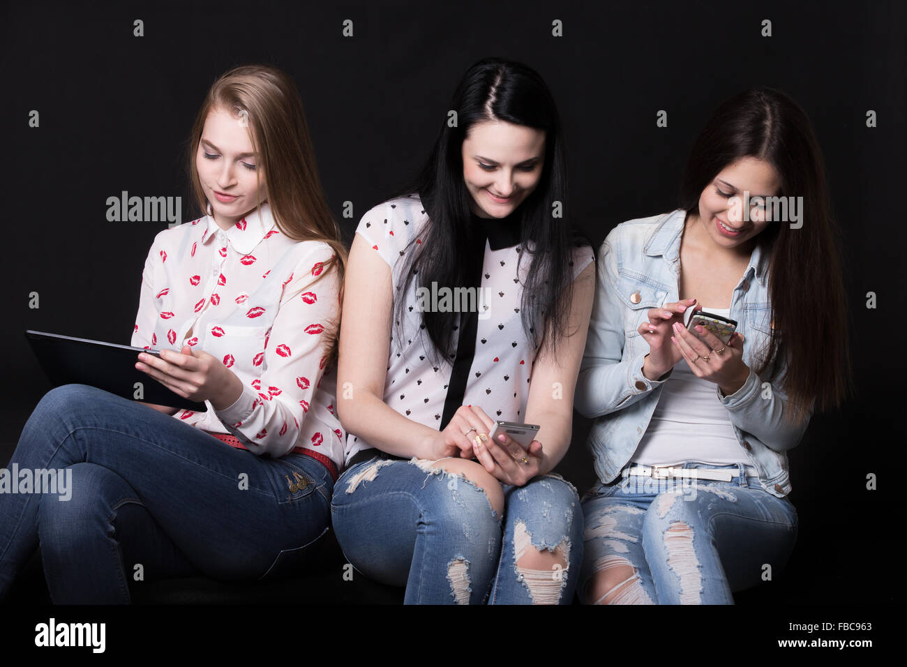 Gruppo di belle ragazze utilizzando diversi dispositivi, seduti insieme ma prestando attenzione solo sui loro telefoni cellulari e ta Foto Stock
