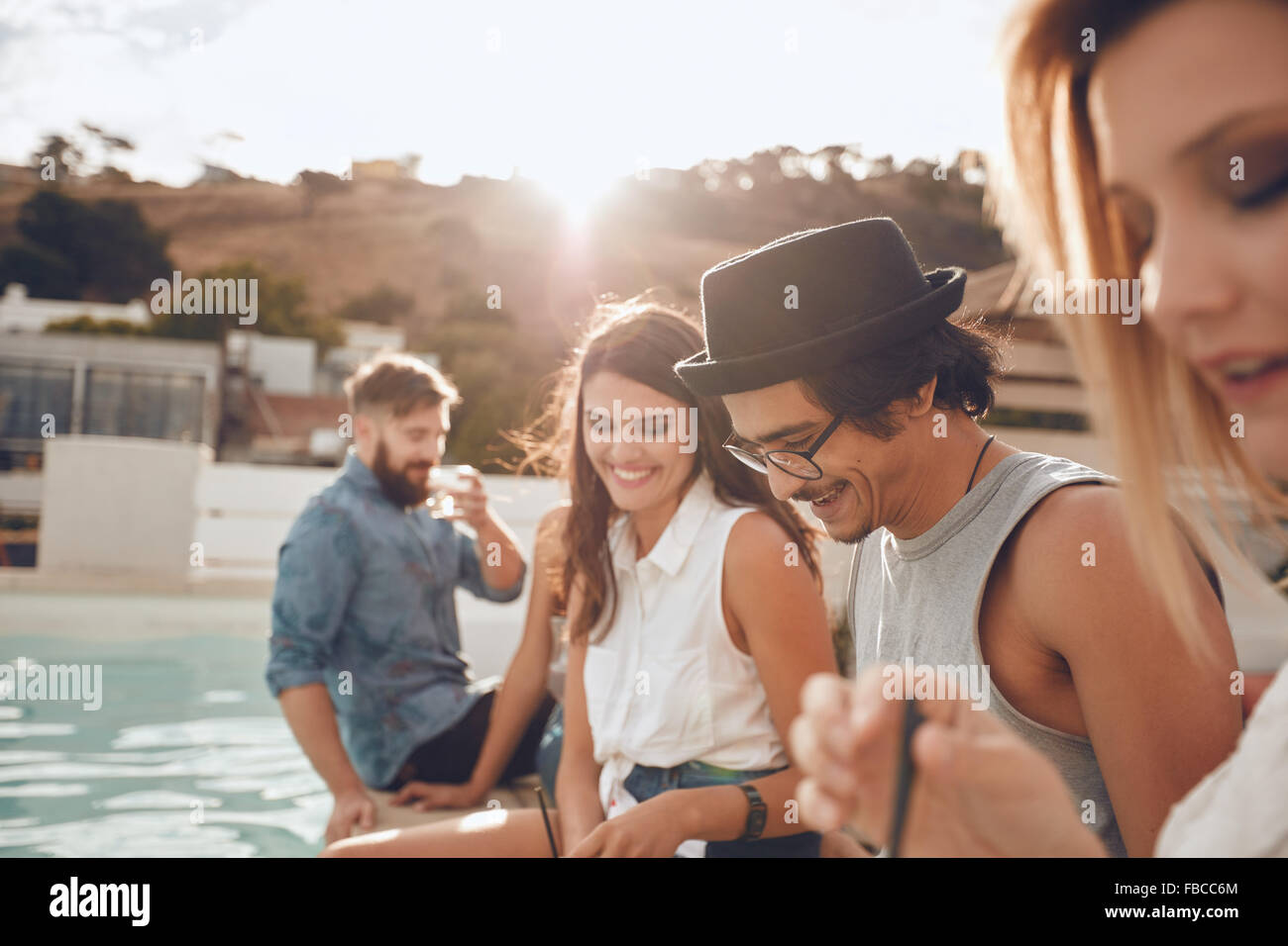 Colpo all'aperto di felice giovane uomo seduto a bordo piscina con i suoi amici di festa. Un gruppo di giovani appeso attorno a una piscina p Foto Stock