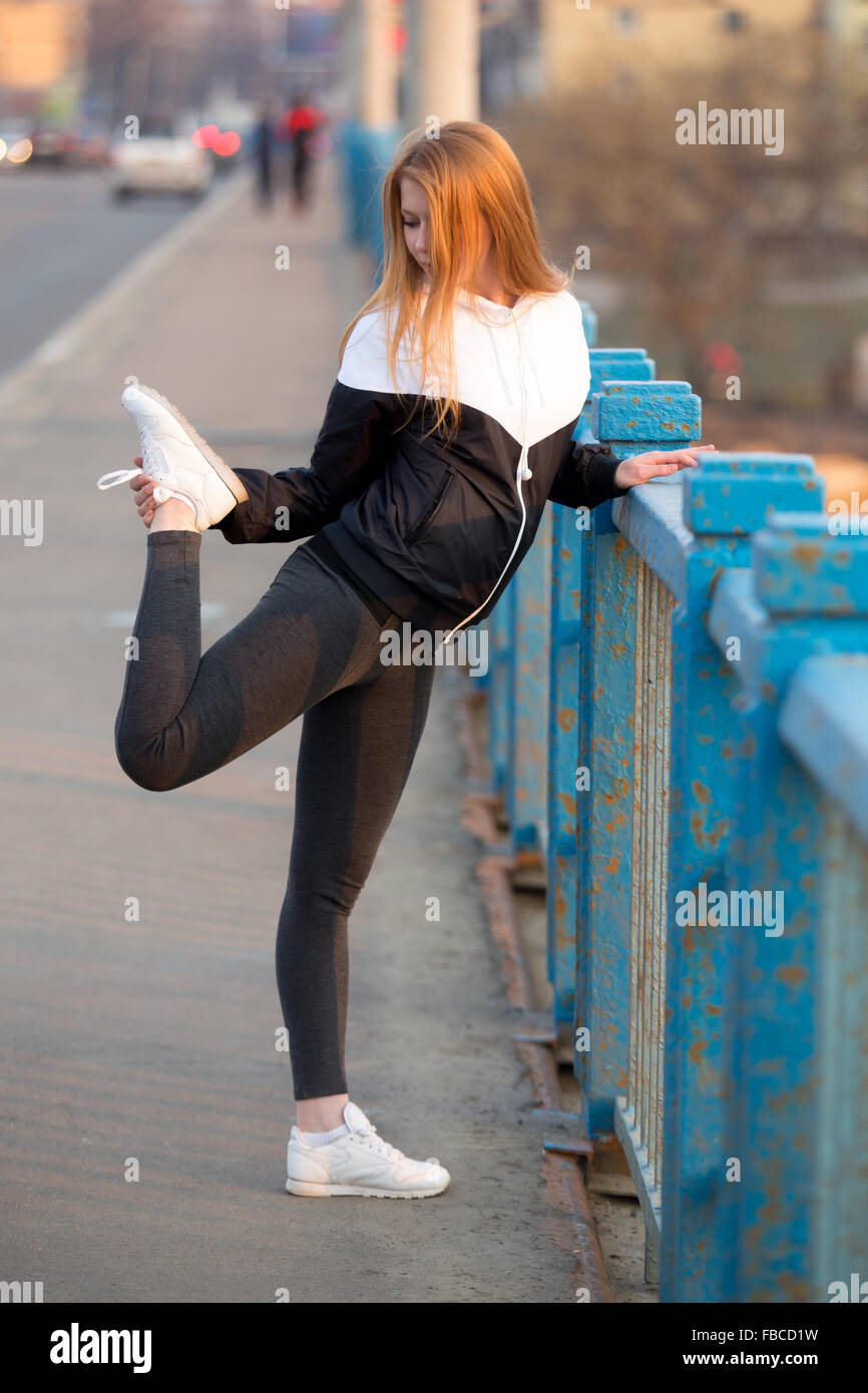 Runner ragazza in abbigliamento sportivo facendo esercizi di stretching per gambe prima di fare jogging sul ponte della città con blue vecchio arrugginito ringhiera, prepari Foto Stock