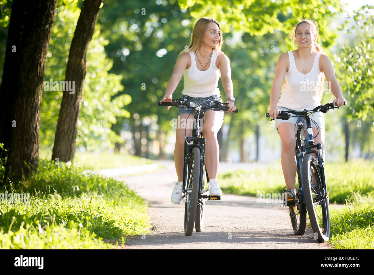Due sorridenti giovani donne fidanzate che indossa jeans corti godono di equitazione biciclette su strada sulla soleggiata giornata estiva, avente conve Foto Stock