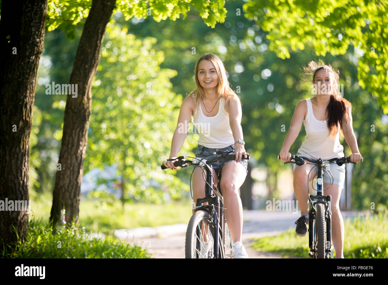 Ritratto di due giovani belle donne allegro fidanzate indossano jeans corti ciclismo su strada sul marciapiede soleggiata giornata estiva, h Foto Stock