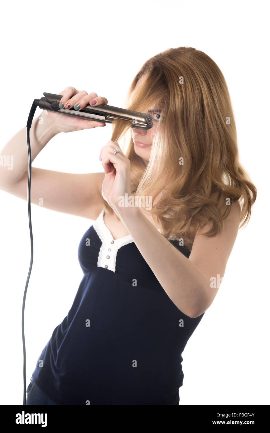 Giovane bella donna bionda la raddrizzatura di suoi lunghi capelli utilizzando ferro elettrico raddrizzatore, studio isolare shot su bianco backgroun Foto Stock