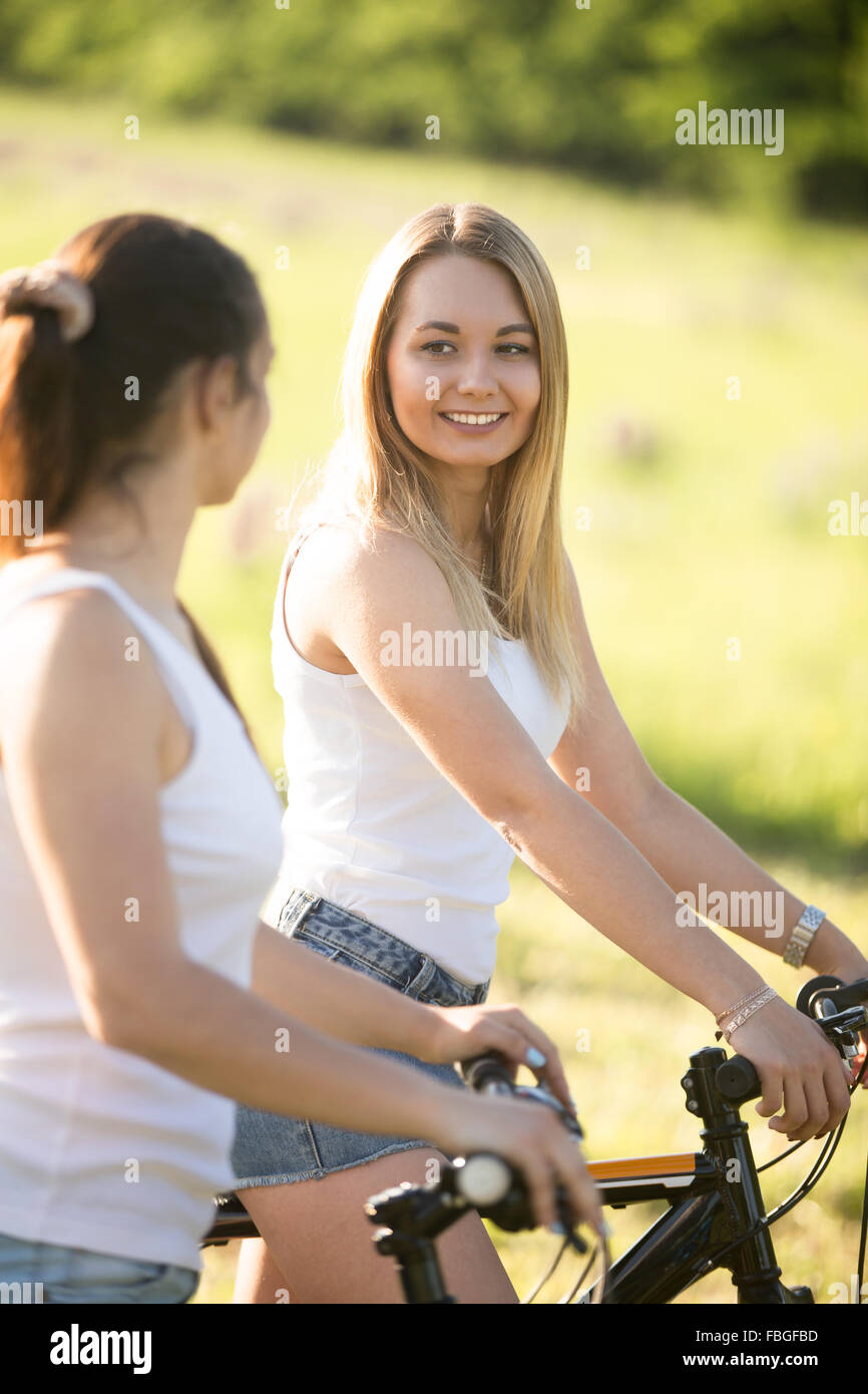 Due giovani belle donne allegro fidanzate indossano jeans corti su biciclette nel parco sulla soleggiata giornata estiva, avente un buon tempo, t Foto Stock