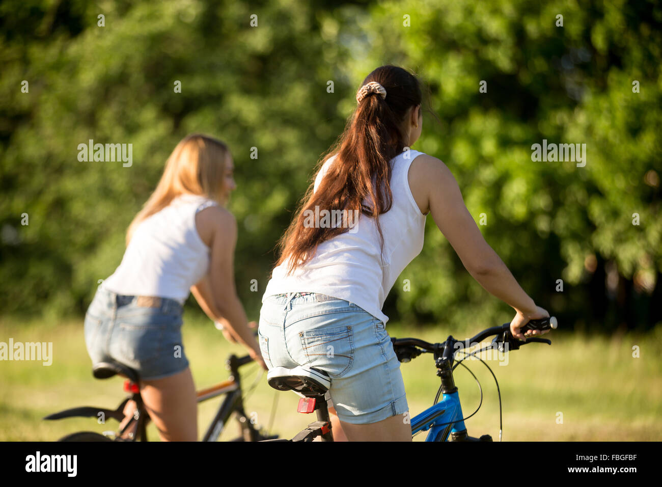 Due giovani donne belle ragazze indossano jeans corti in bicicletta nel parco sulla soleggiata giornata estiva, vista posteriore Foto Stock