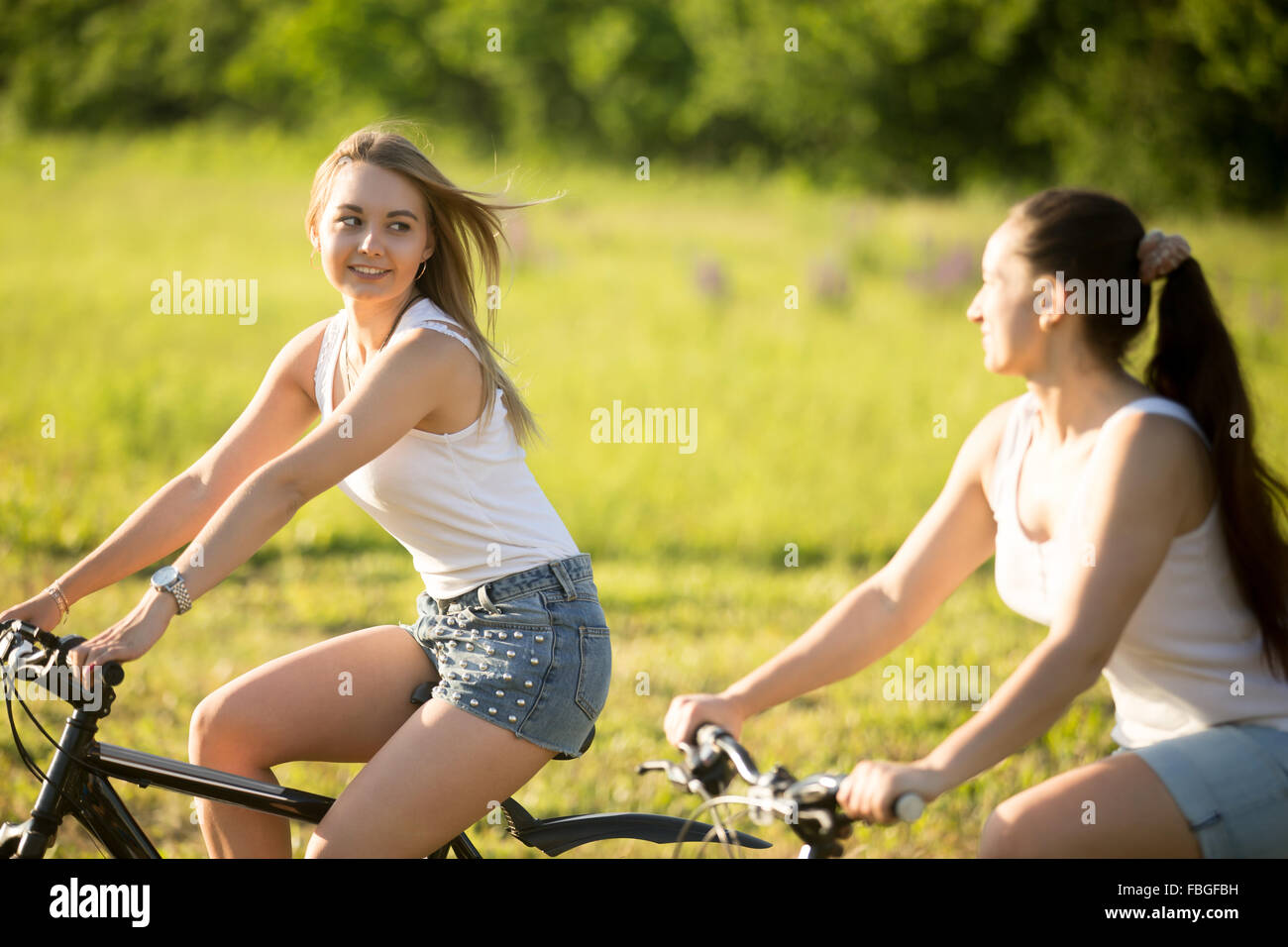 Due simpatici giovani sorridenti donne belle ragazze indossano jeans corti moto di equitazione nel parco in condizioni di luce solare intensa in estate Foto Stock