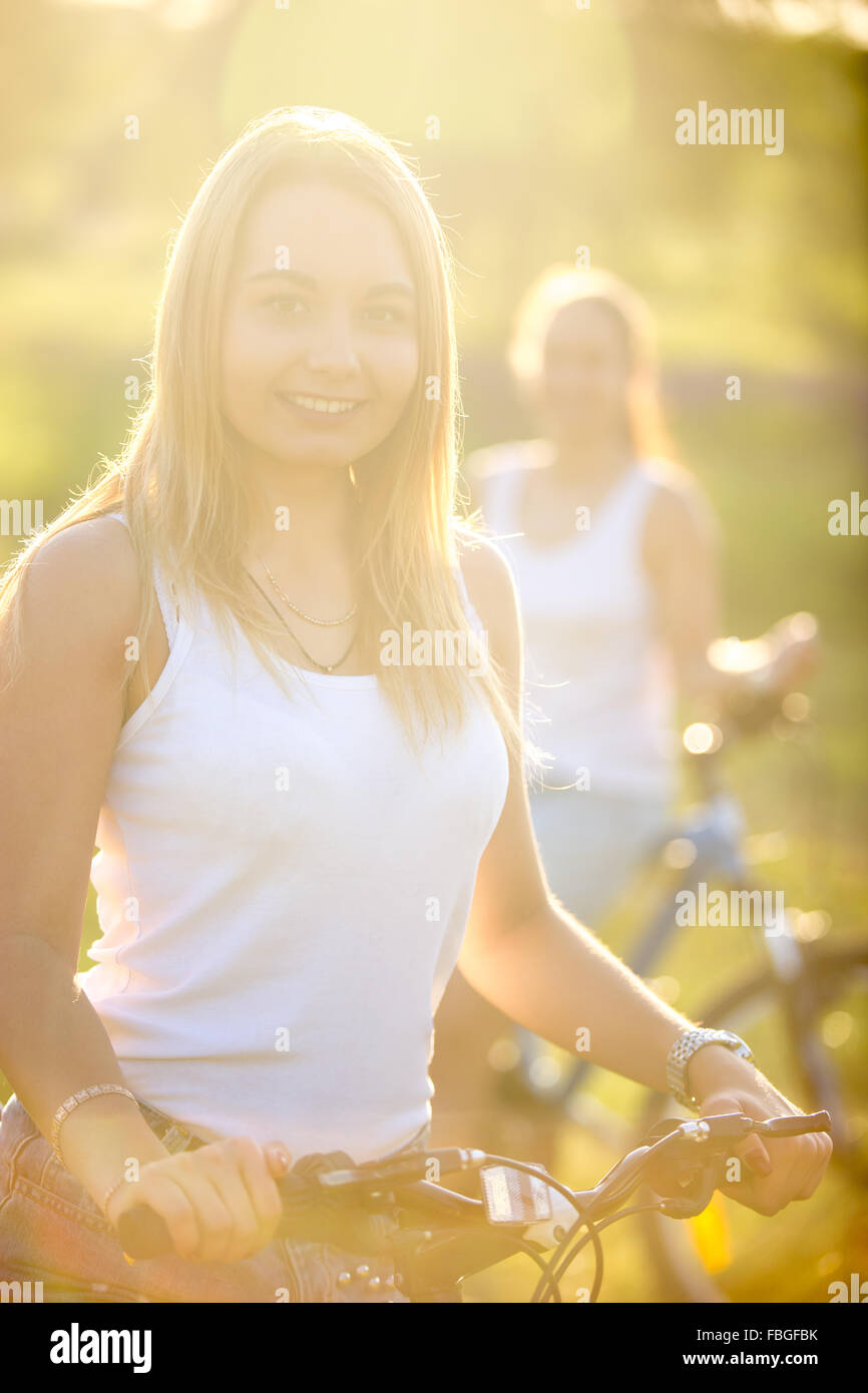 Due allegro bella ciclista teenage fidanzate indossando bianco casual canotte in piedi con le biciclette nel parco in bright sun Foto Stock