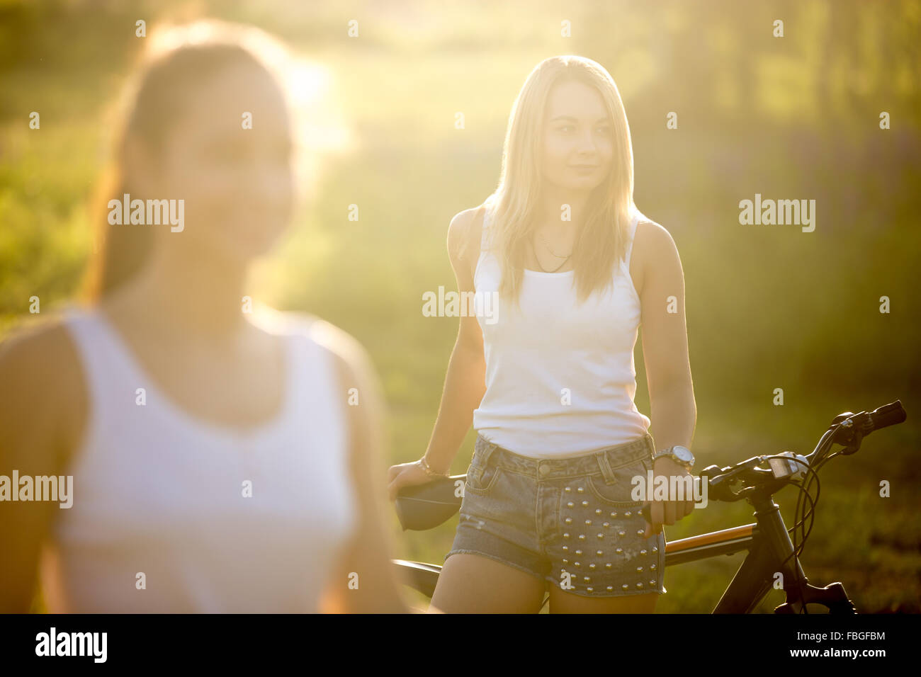 Due amiche attraente indossando bianco casual canotte e jeans corti in piedi con la moto in campagna parco sul sole luminoso Foto Stock