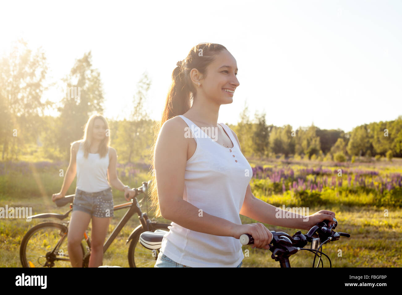 Due belle ragazze con le biciclette indossando bianco casual canotte e gli shorts in denim in piedi in campagna parco sulla luminosa s Foto Stock