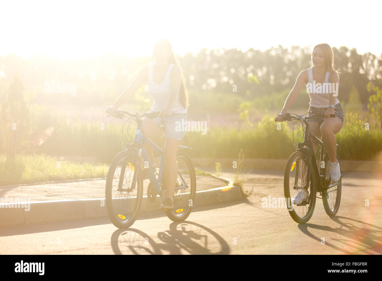 Due belle ragazze adolescenti equitazione biciclette su park via indossando bianco casual canotte e jeans corti sul luminoso estate da Foto Stock
