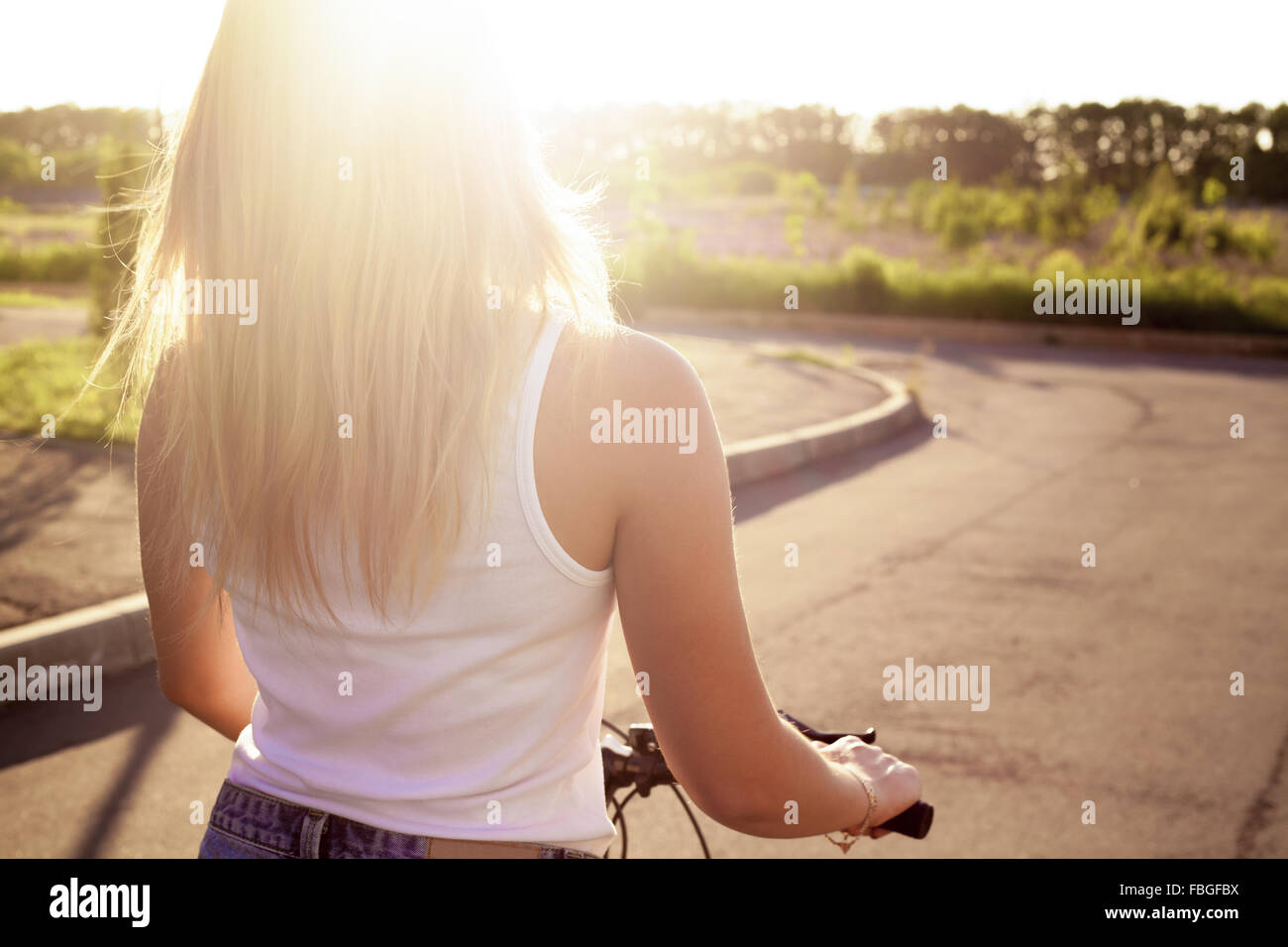 Bella ragazza adolescente riding bike indossa casual serbatoio bianco top e jeans corti sul parco sulla strada illuminata dal sole giornata estiva, indietro Foto Stock