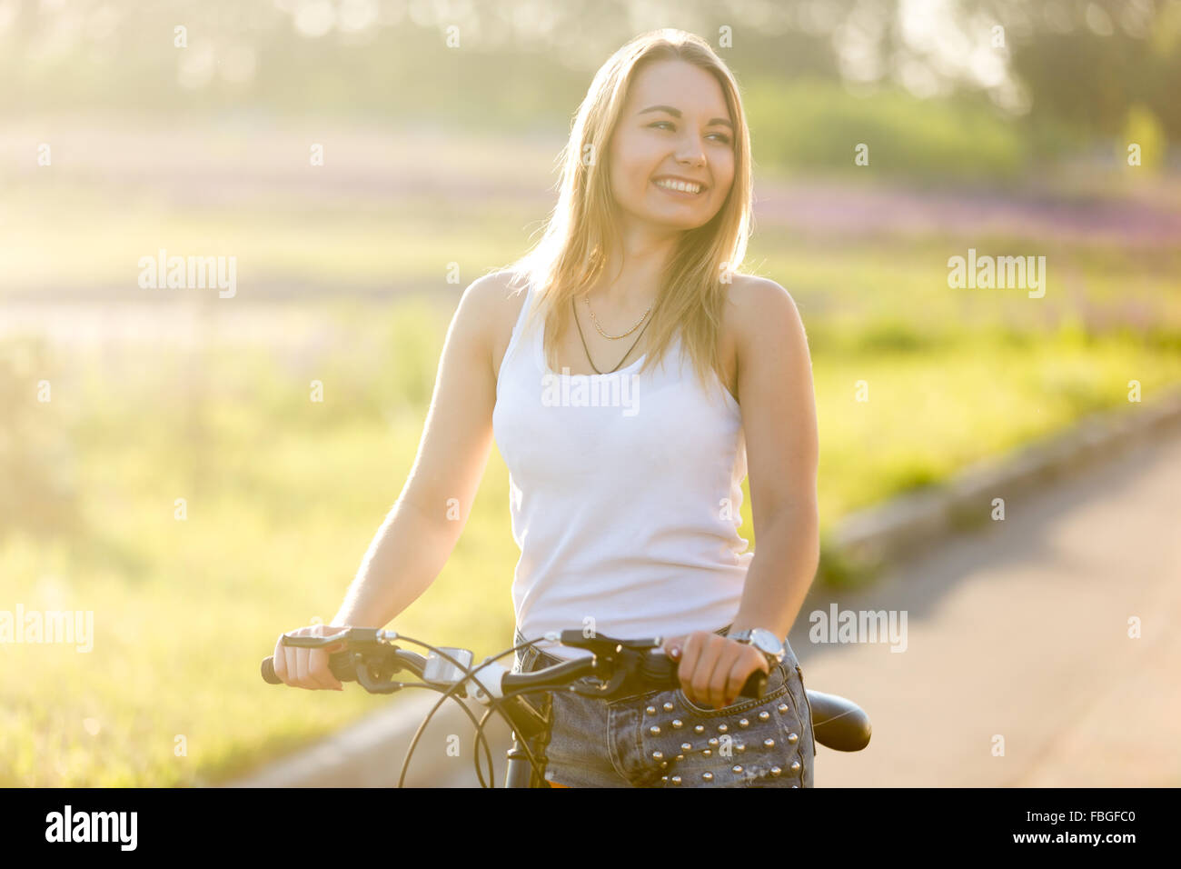 Ritratto di ridere bella giovane donna seduta su mountain bike indossa casual serbatoio bianco top e jeans su park road a Brig Foto Stock