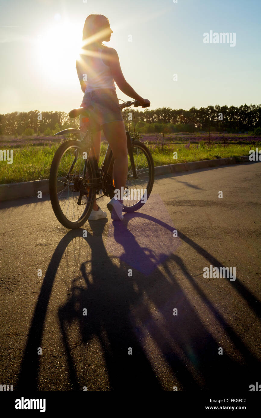 Vista posteriore della silhouette sportiva bella giovane donna sulla bici indossando casual serbatoio bianco top e jeans corti sul giorno di estate, sole s Foto Stock