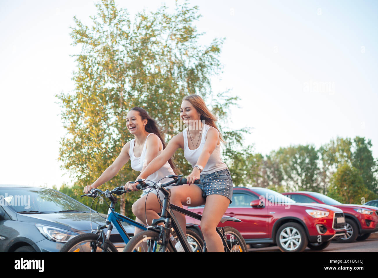 Due lieti di ridere bella bionda e bruna di donne giovani amici di equitazione in giocoso bike race insieme a fianco di automobili parcheggiate outd Foto Stock
