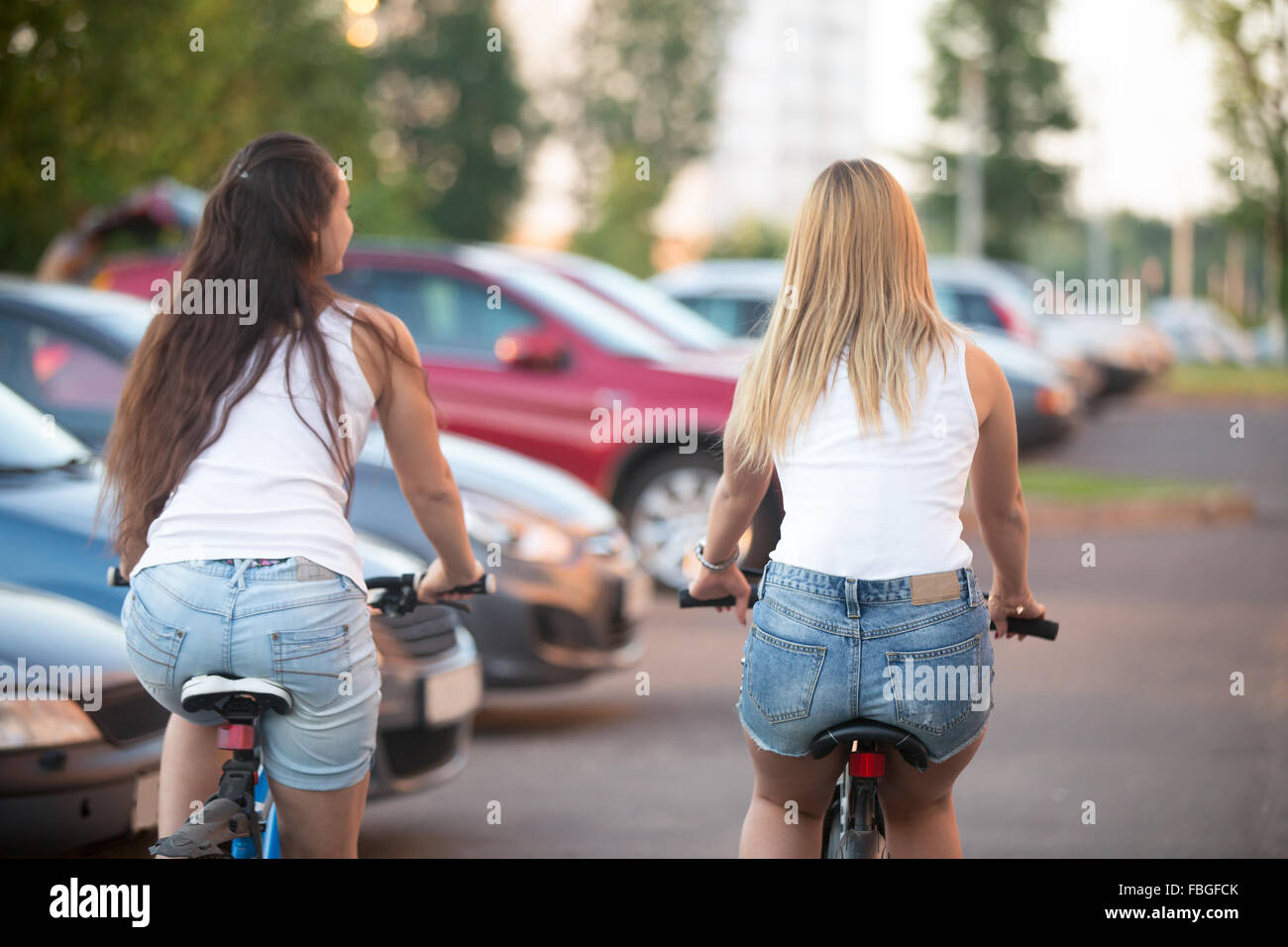 Due bellissime bionde e bruna di donne giovani amici equitazione biciclette in città in estate a fianco di una vettura parcheggiata all'aperto a residenti Foto Stock