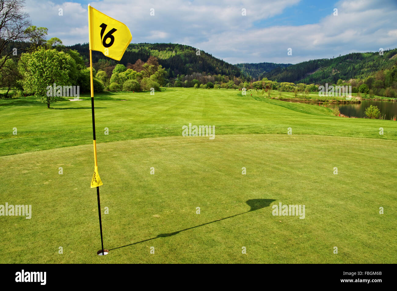 Bandiera gialla nel foro sul verde del campo da golf, fairway accanto ad un laghetto, delle montagne boscose in background Foto Stock