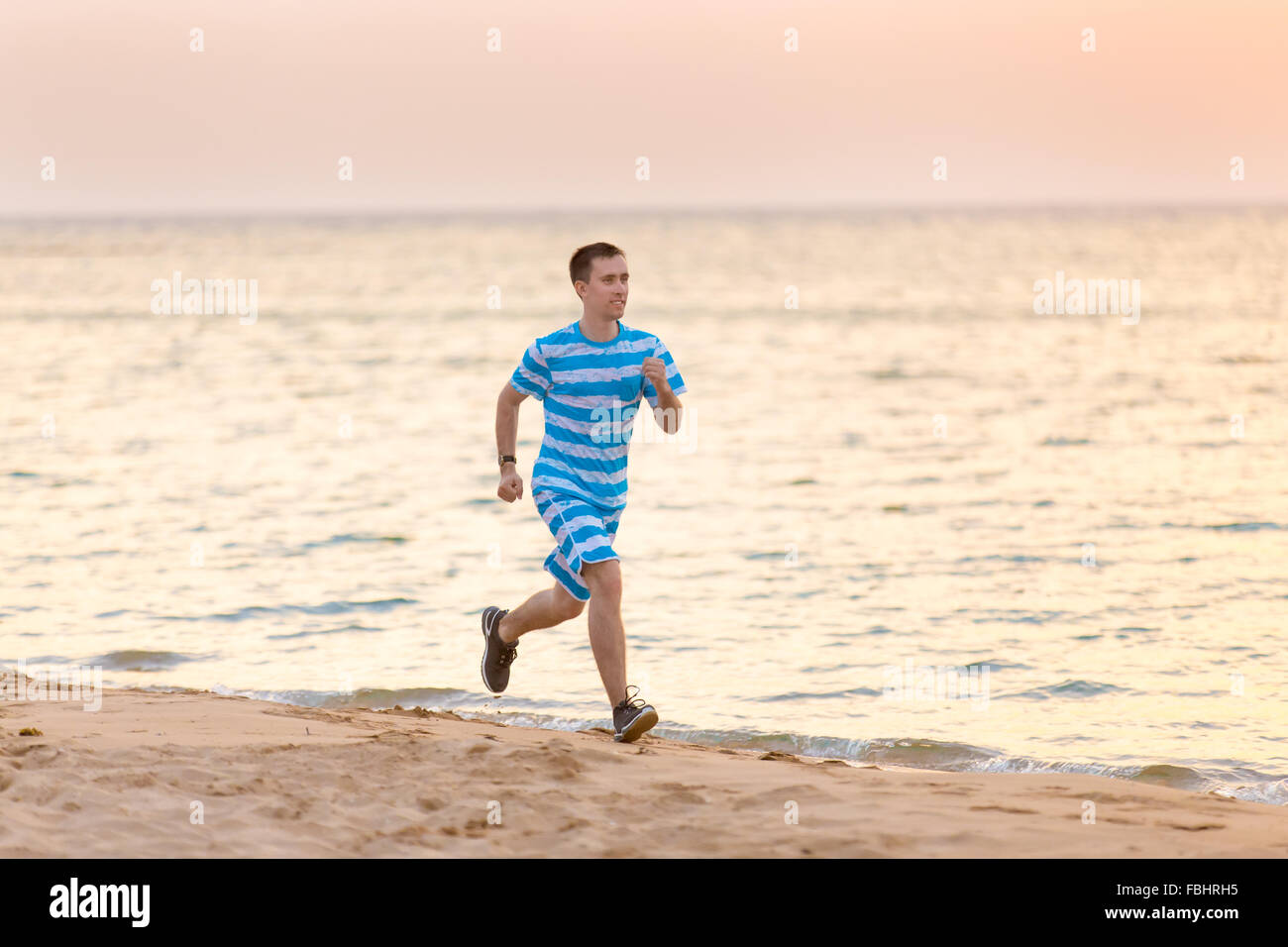 Uno stile di vita sano: bello allegro giovane uomo che lavora al di fuori, corre veloce sulla costa di sabbia al tramonto sul tramonto o l'alba Foto Stock