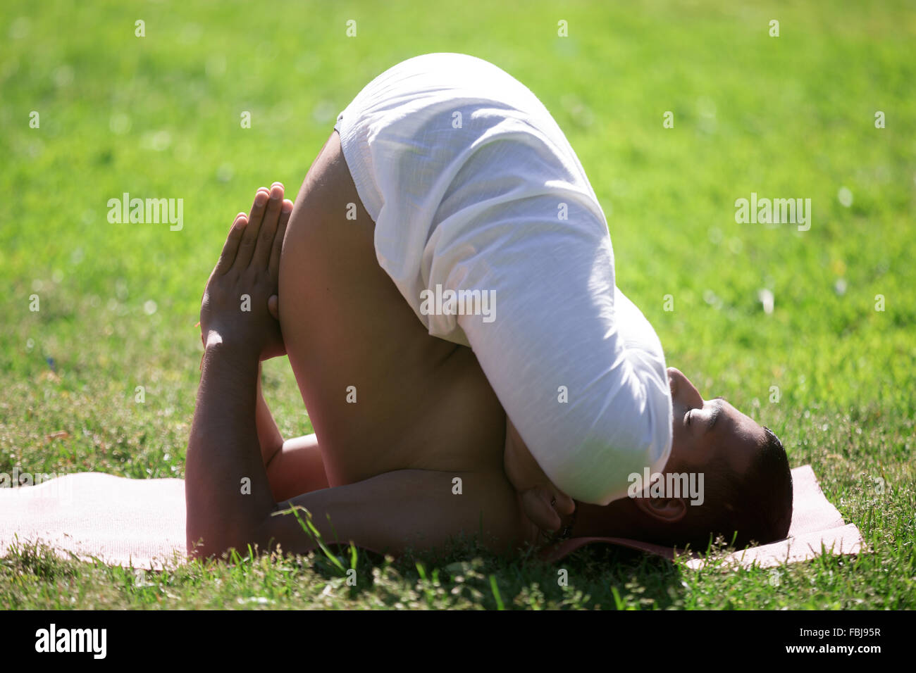 Indiano attraente giovane uomo che lavora fuori del tappetino sul prato nel parco, facendo pindasana in sarvangasana, variazione della postura embrionale, fu Foto Stock