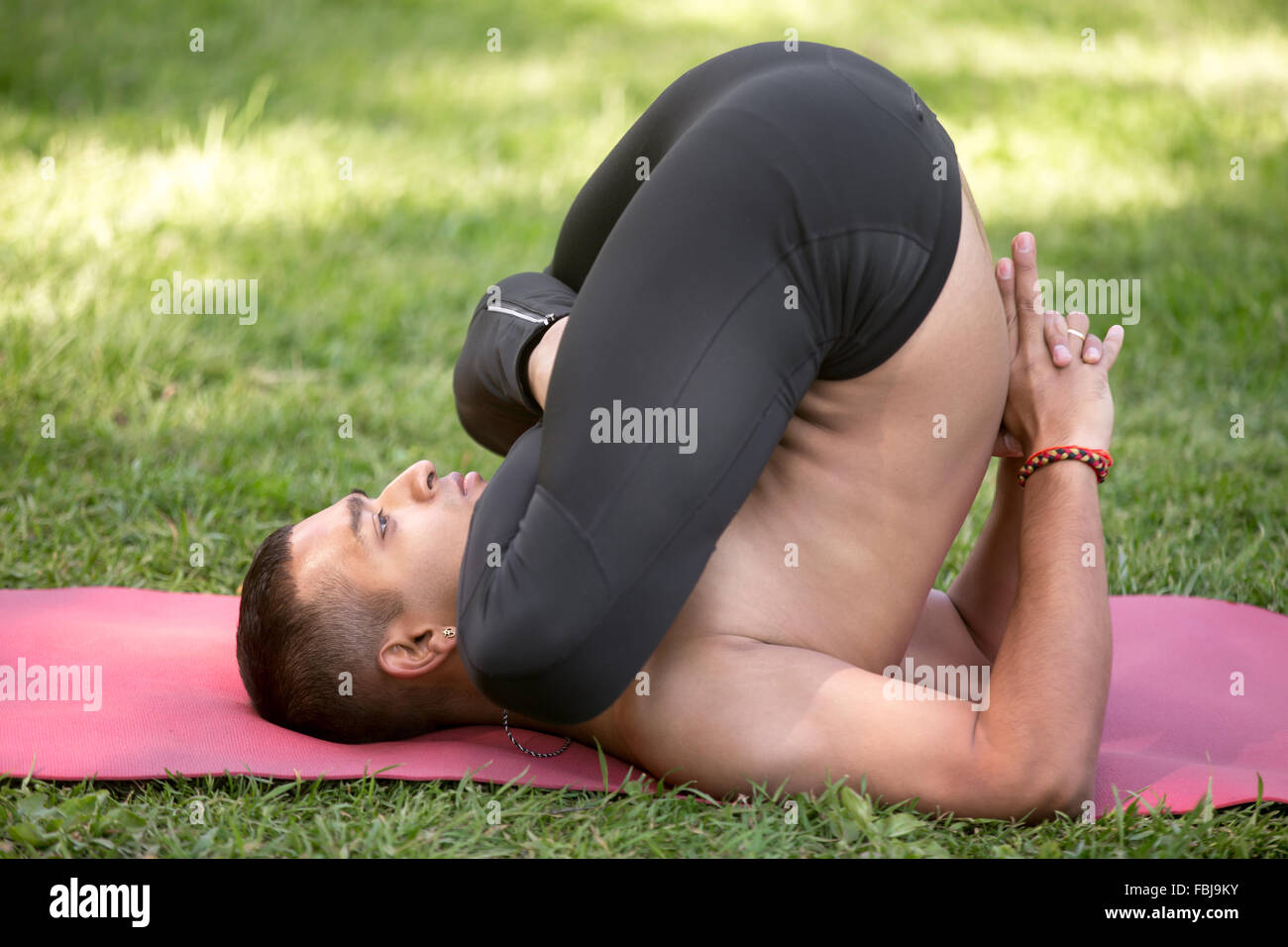 Indiano bello giovane uomo che lavora fuori del tappetino sul prato nel parco, facendo pindasana in sarvangasana, variazione della postura embrionale, pieno Foto Stock