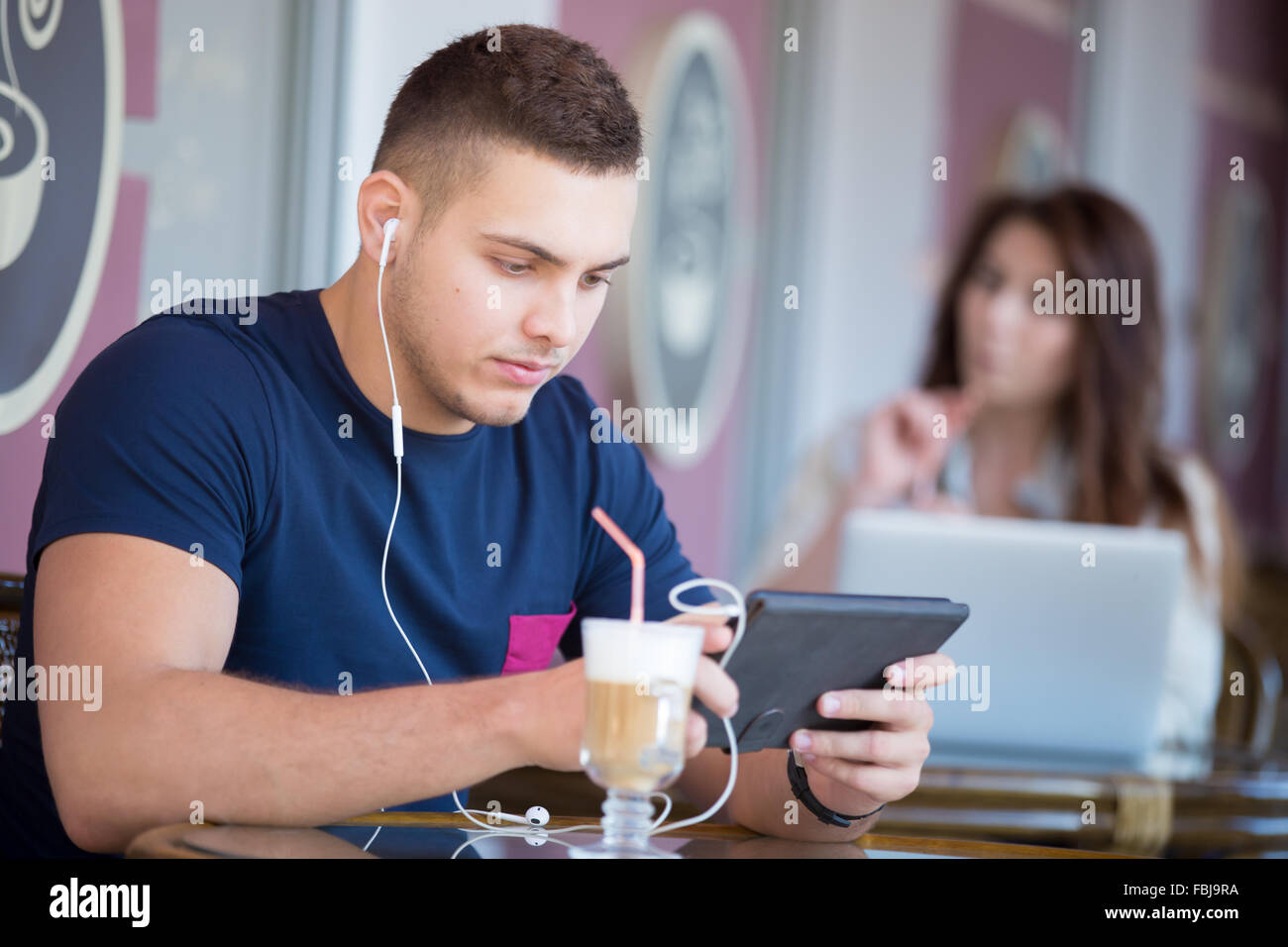 Ritratto di grave bel giovane con auricolari, utilizzando la tavoletta, guardando lo schermo, seduta in street cafe con vetro di Foto Stock