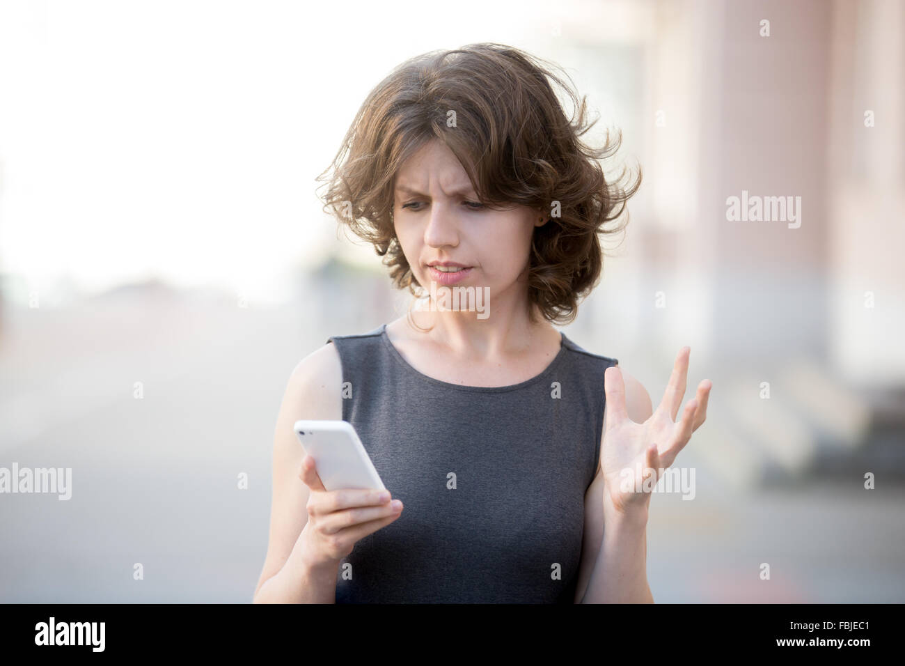 Ritratto di giovane donna tenendo il cellulare in mani sulla strada in estate, guardando lo schermo con espressione irritata, fatta Foto Stock