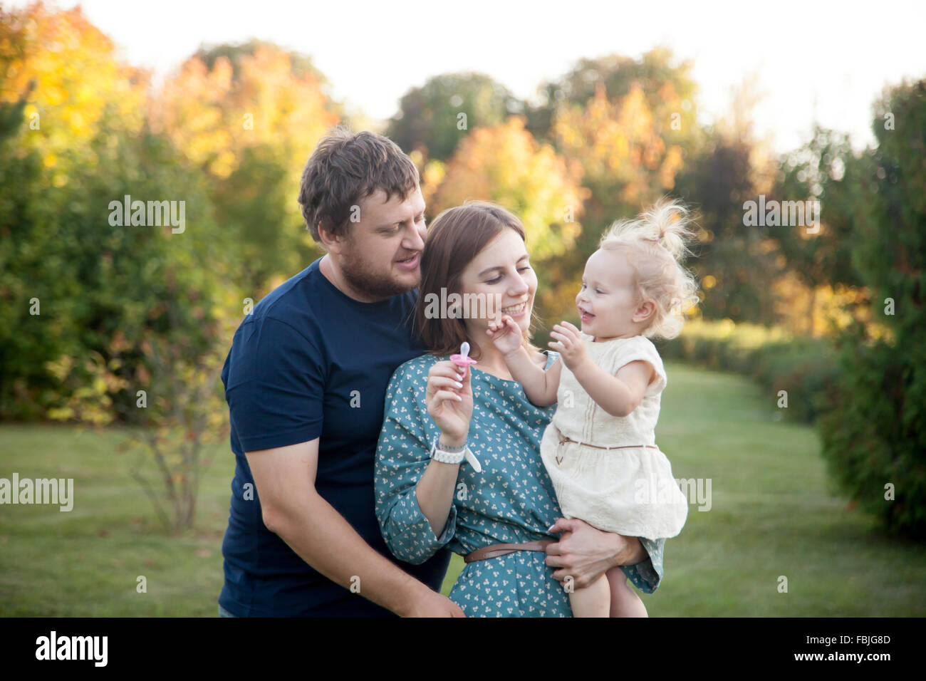 Ritratto di felice bella famiglia di tre persone che camminano nel parco in autunno. Mamma e papà bambino portando la figlia in abito bianco, Foto Stock