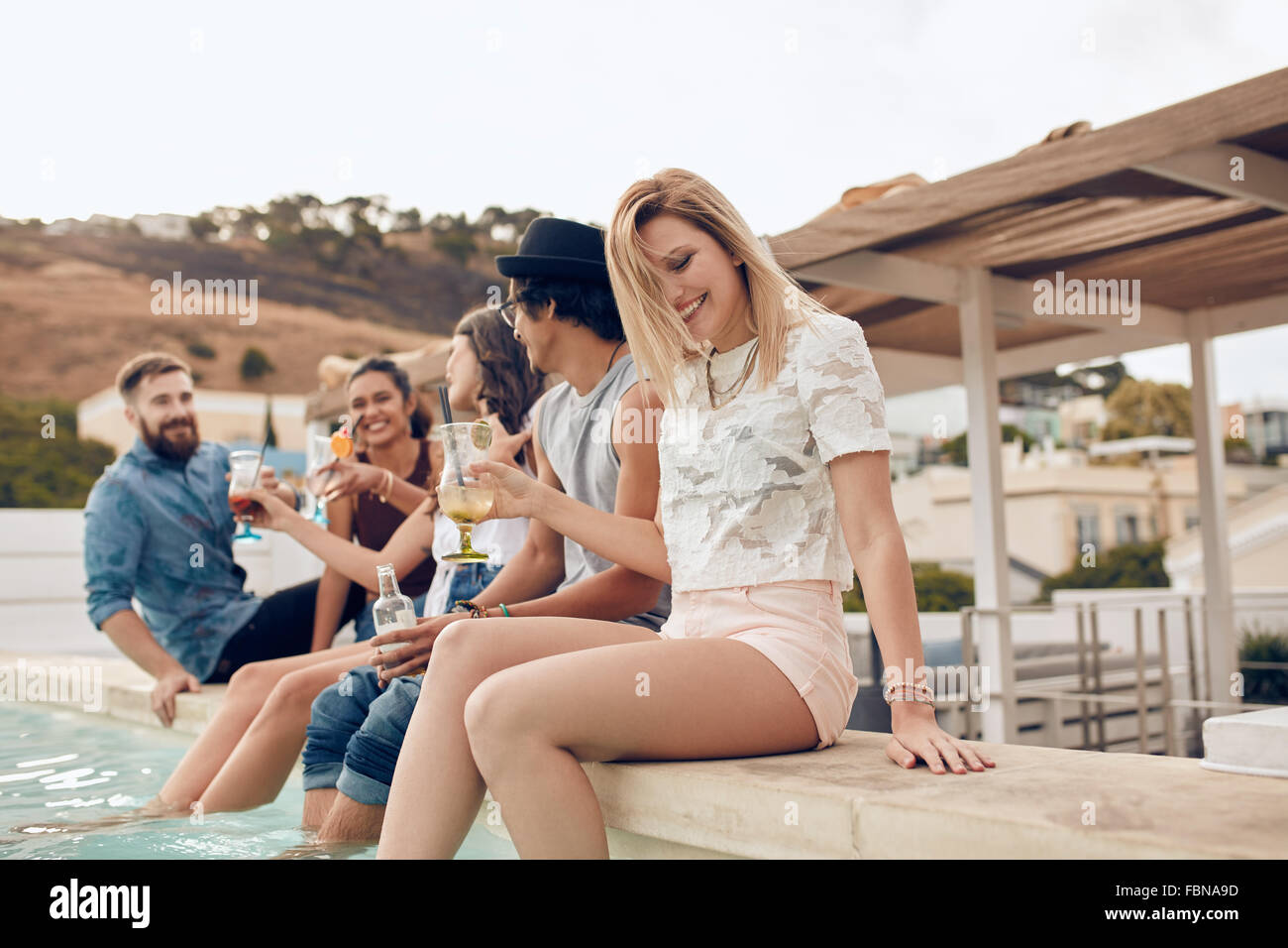 Colpo all'aperto di felice giovani aventi parti. Giovani uomini e donne seduta sul bordo della piscina con i piedi in acqua Foto Stock