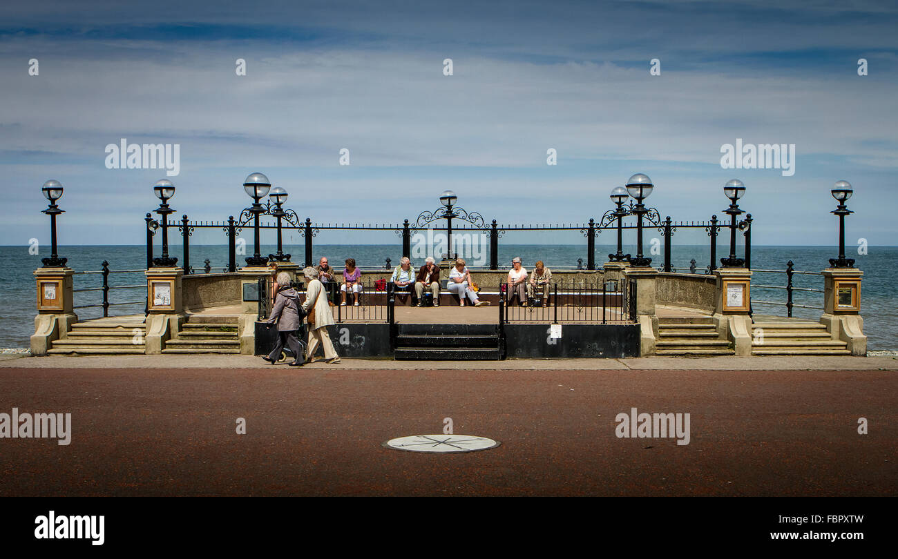Gli anziani seduti sui sedili e oltrepassando il bandstand lungo il lungomare di Llandudno Foto Stock