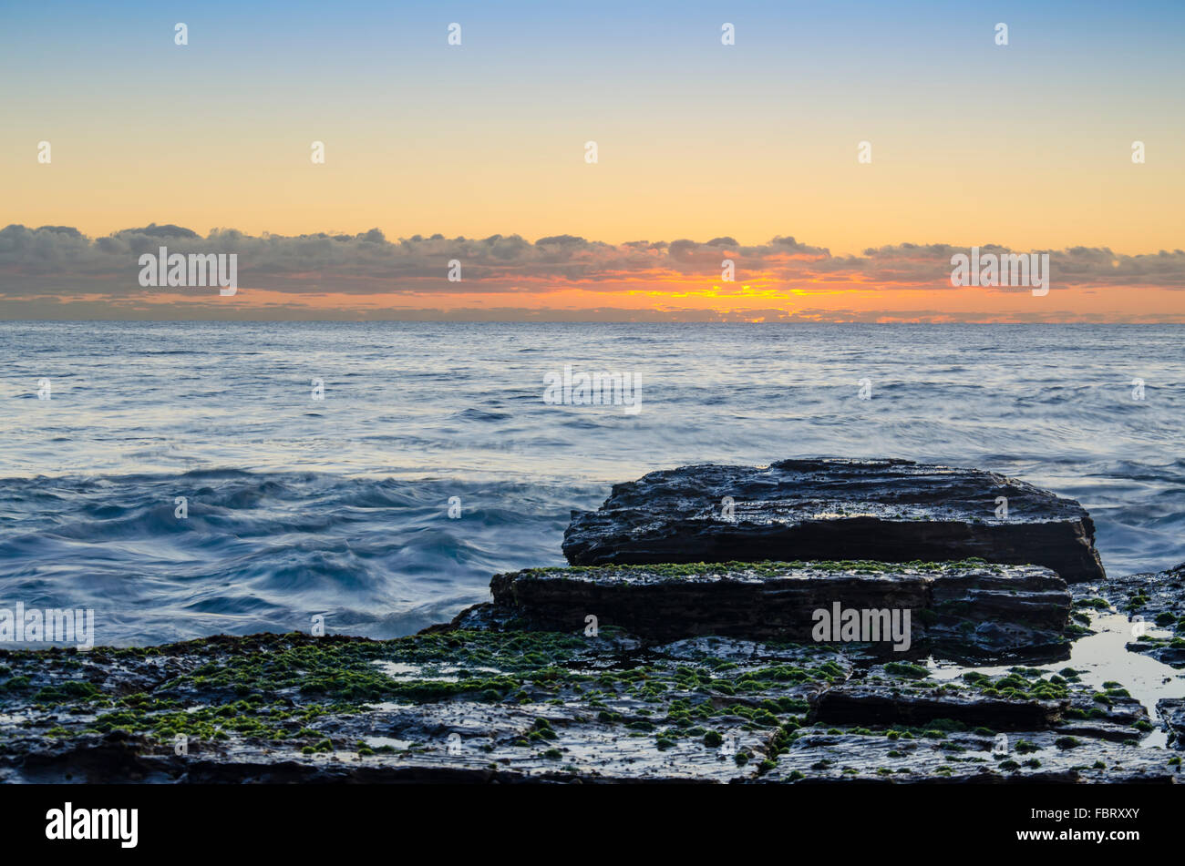 Con un'alba dorata e una linea di nubi di cumulo basso sullo sfondo, un basso rigonfiamento porta le onde verso una sporgenza di roccia sulla costa orientale dell'Australia Foto Stock