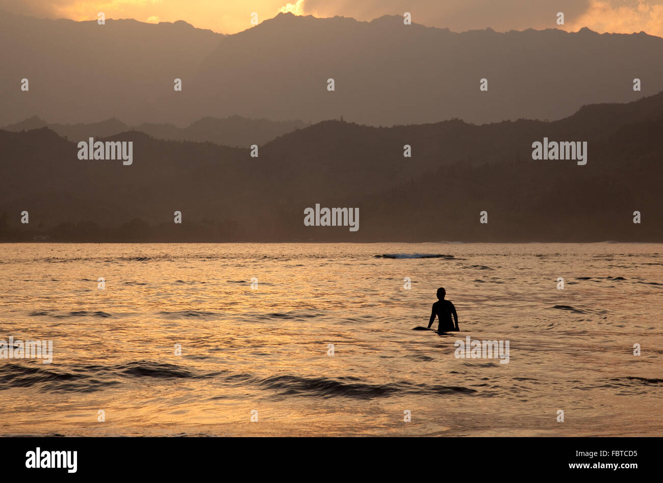 Tramonto dietro Na Pali di montagna con una silhouette del surfer in primo piano e il sole riflette sull'oceano Foto Stock