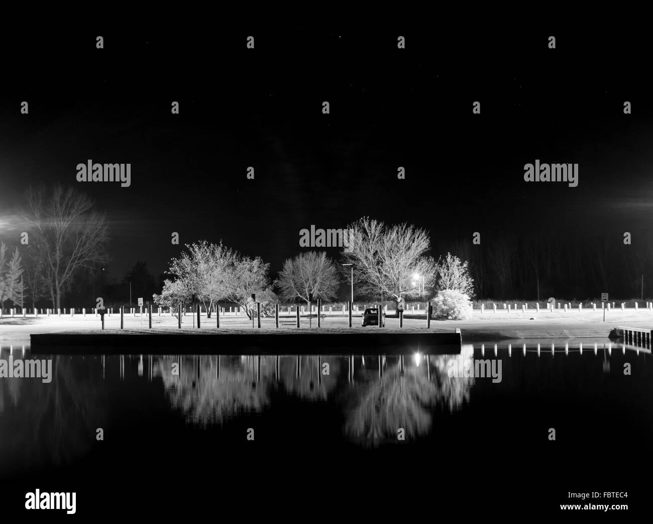 Una barca il lancio di notte è illuminato da luci di strada facendo in modo che gli alberi sfrondato per candelette di bianco, in contrasto con il nero come specchio di acqua Foto Stock