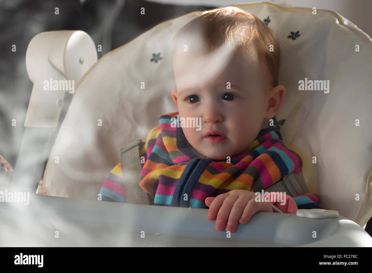 Un anno di età baby ragazza sedeva in un seggiolone con il fumo di sigaretta in primo piano suggerendo il fumo passivo Rischio Foto Stock