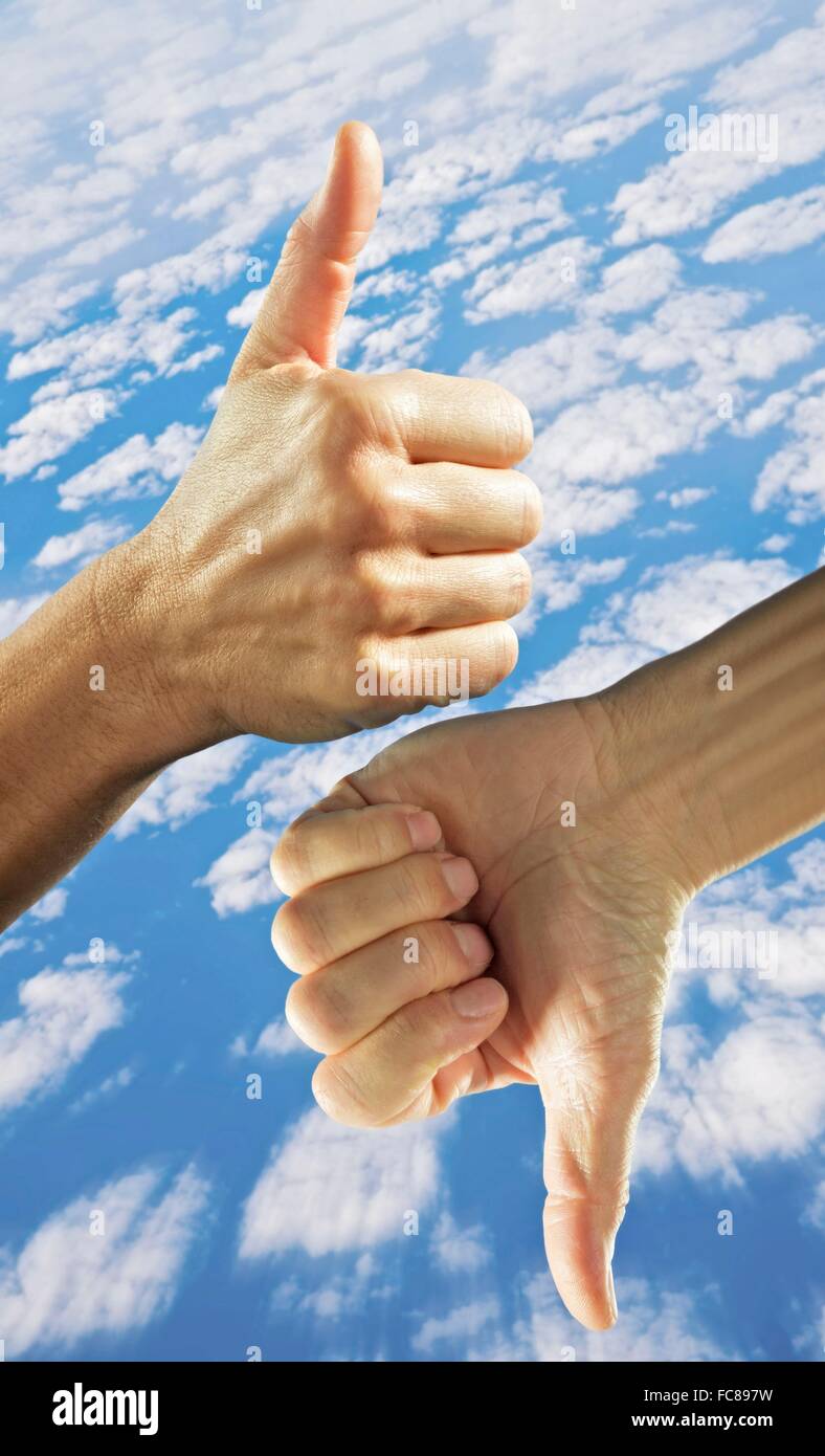 Una mano con il pollice in alto e il pollice verso il basso contro il cielo nuvoloso. Foto Stock