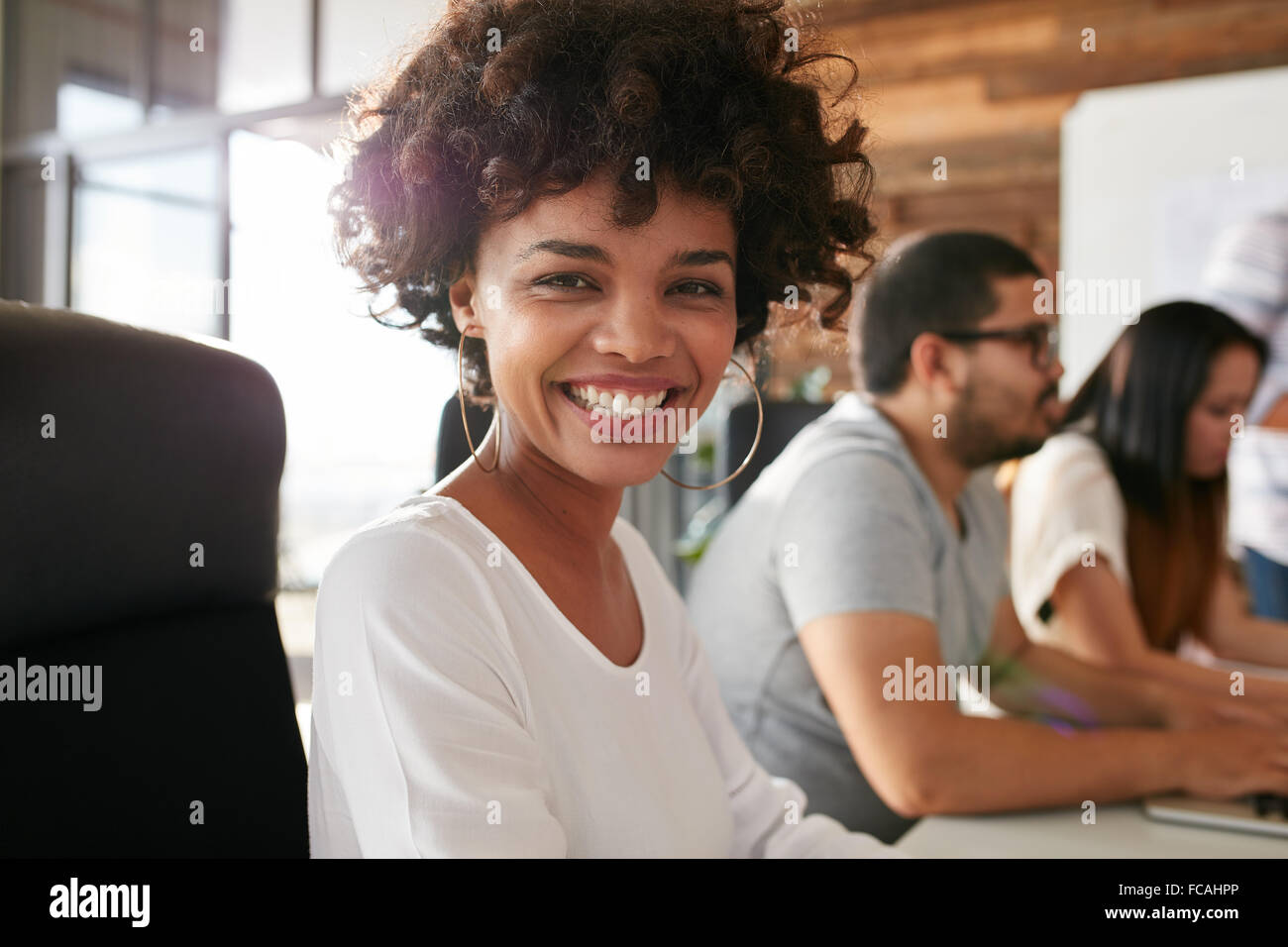Closeup Ritratto di Allegro giovane donna africana seduti nella sala conferenze con i colleghi di lavoro in background. Riunione di team Creative Foto Stock