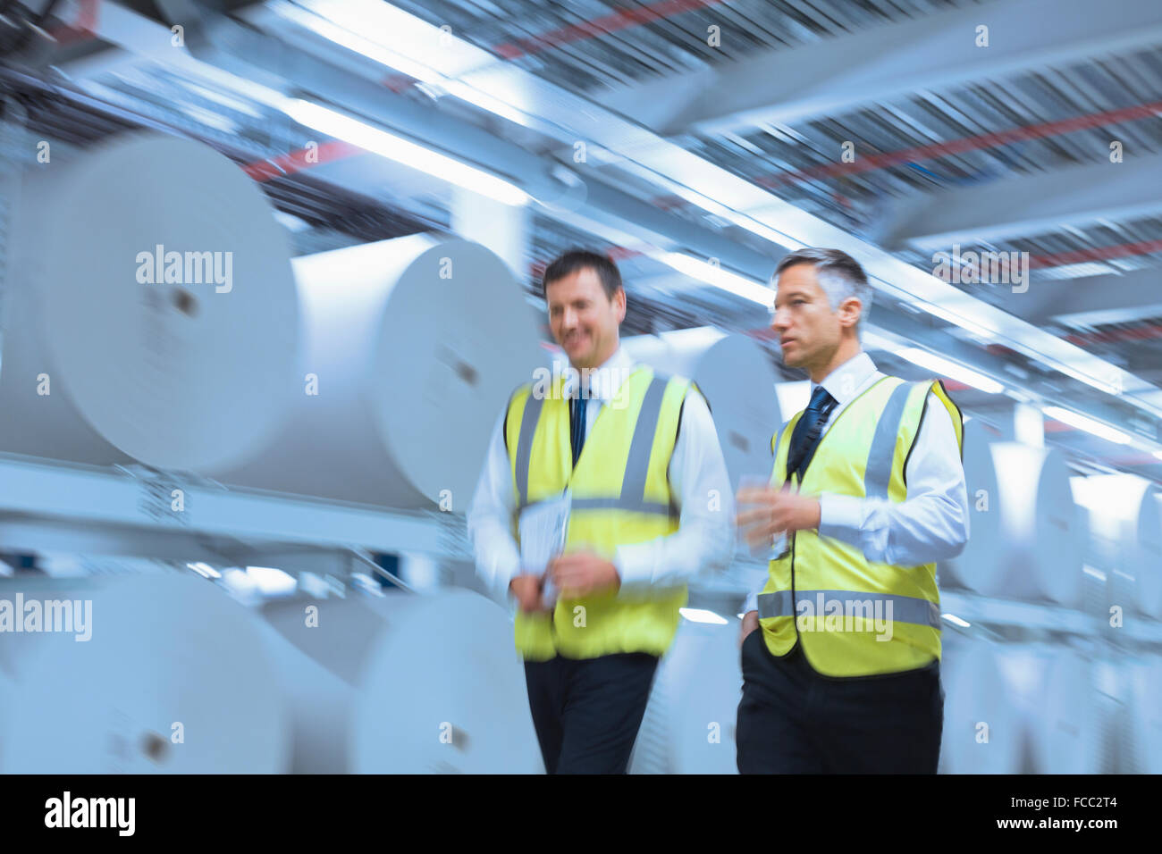 Uomini d'affari in abbigliamento riflettivo a piedi lungo i grandi bobine di carta in impianto di stampa Foto Stock
