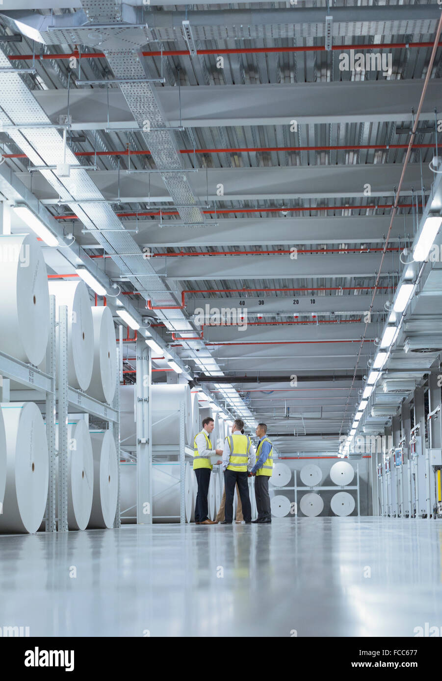 Lavoratori in abbigliamento riflettivo parlando vicino a grandi bobine di carta in impianto di stampa Foto Stock
